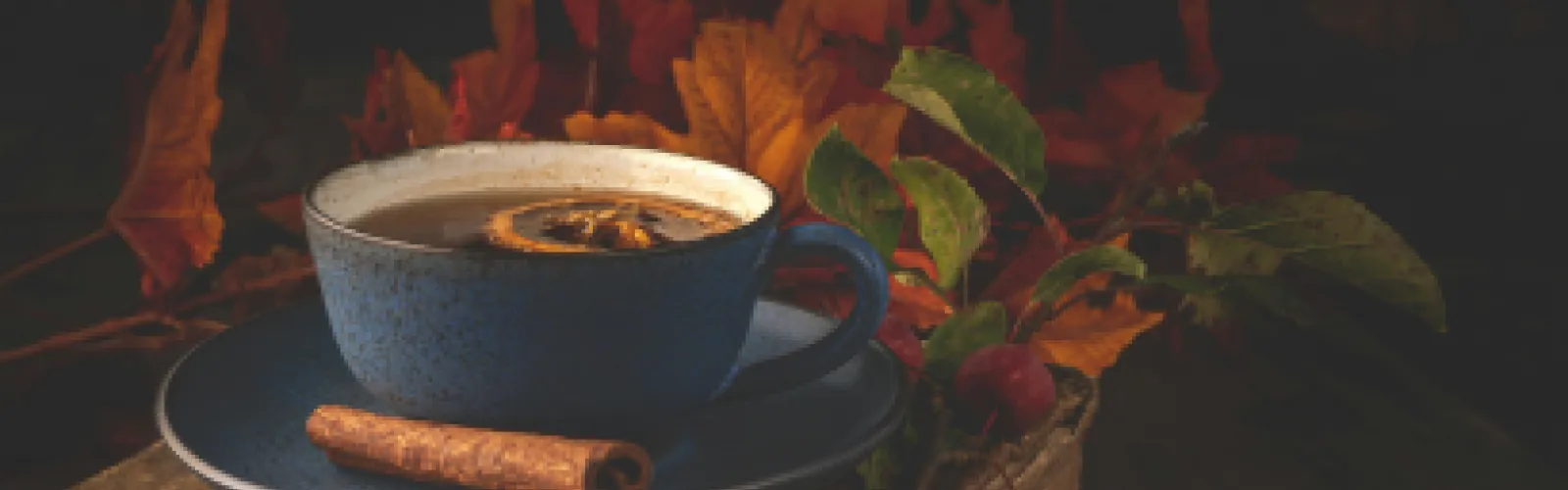 A blue cup of tea with lemon and a cinnamon stick on a saucer set against a backdrop of vibrant autumn leaves.